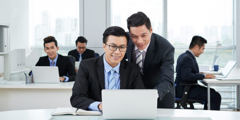 Busy open plan office: handsome Asian businessman standing at desk and explaining how to operate computer program to his subordinate
